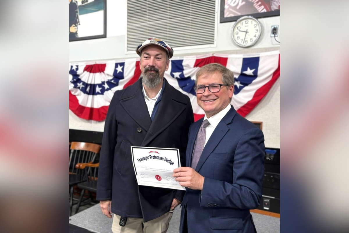 Jason Osborne (left) and Grover Norquist (right) hold a certificate titled “Taxpayer Protection Pledge.” Osborne wears a patterned flat cap, dark coat, and has a gray beard. Norquist wears glasses and a blue suit. American flag bunting hangs on the wall behind them.