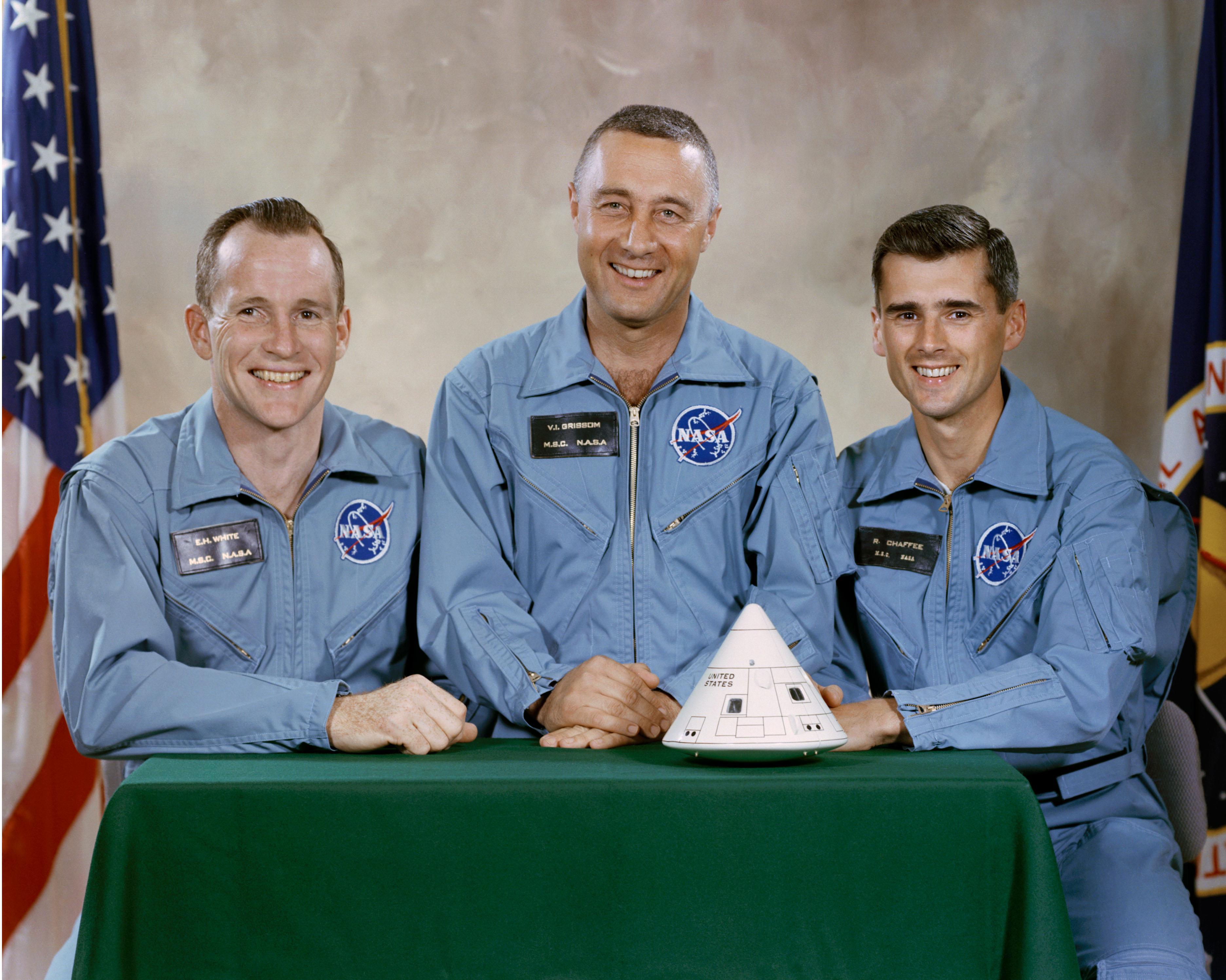 The Apollo 1 crew — Ed White, Gus Grissom and Roger Chaffee — in blue NASA flight suits behind a model of the Apollo command module.