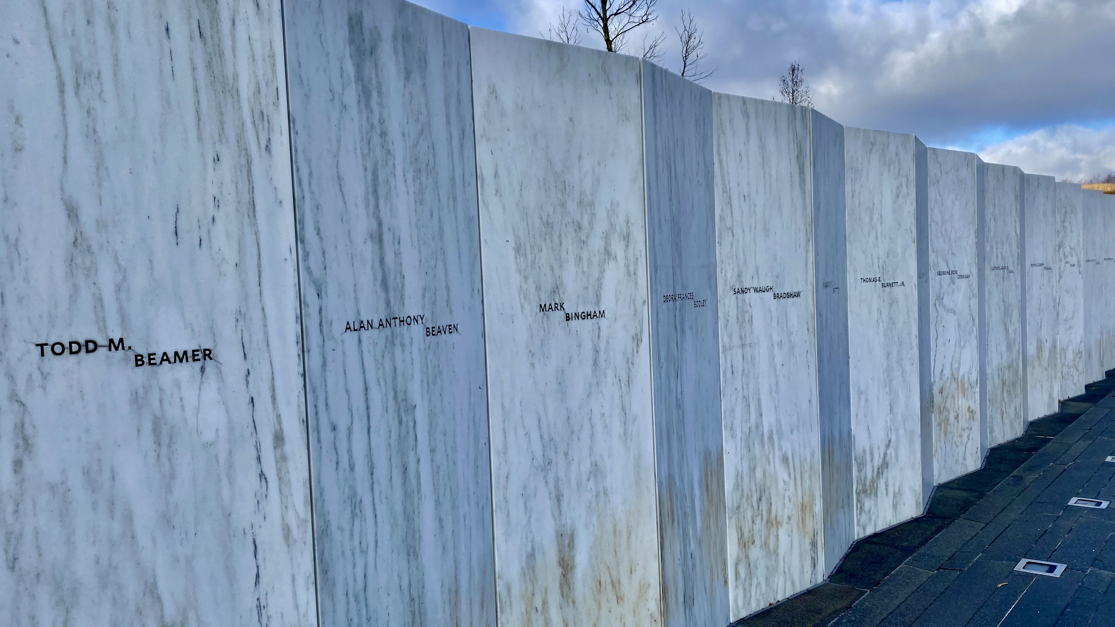 The Wall of Names at the Flight 93 National Memorial: a row of angled white marble panels, each carved with the name of a passenger or crew member. Todd M. Beamer, Alan Anthony Beaven, Mark Bingham, and Deora Frances Bodley are legible in the foreground, with the rest of the wall receding toward a bare treeline under a winter sky.