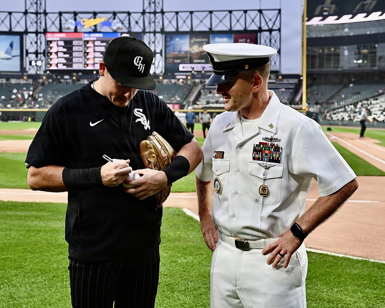 White Sox outfielder Sam Antonacci, in a black jersey and cap, signs a baseball for a smiling U.S. Navy officer in dress whites on the field at Rate Field, with the stands and scoreboard behind him.