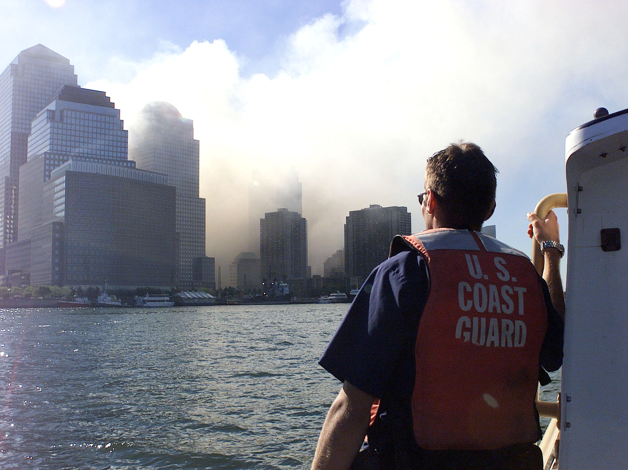 A U.S. Coast Guard crewman stands at the rail of a boat in New York Harbor, facing the Lower Manhattan skyline, where a wall of pale smoke rises from the collapsed World Trade Center and blots out the buildings behind it.