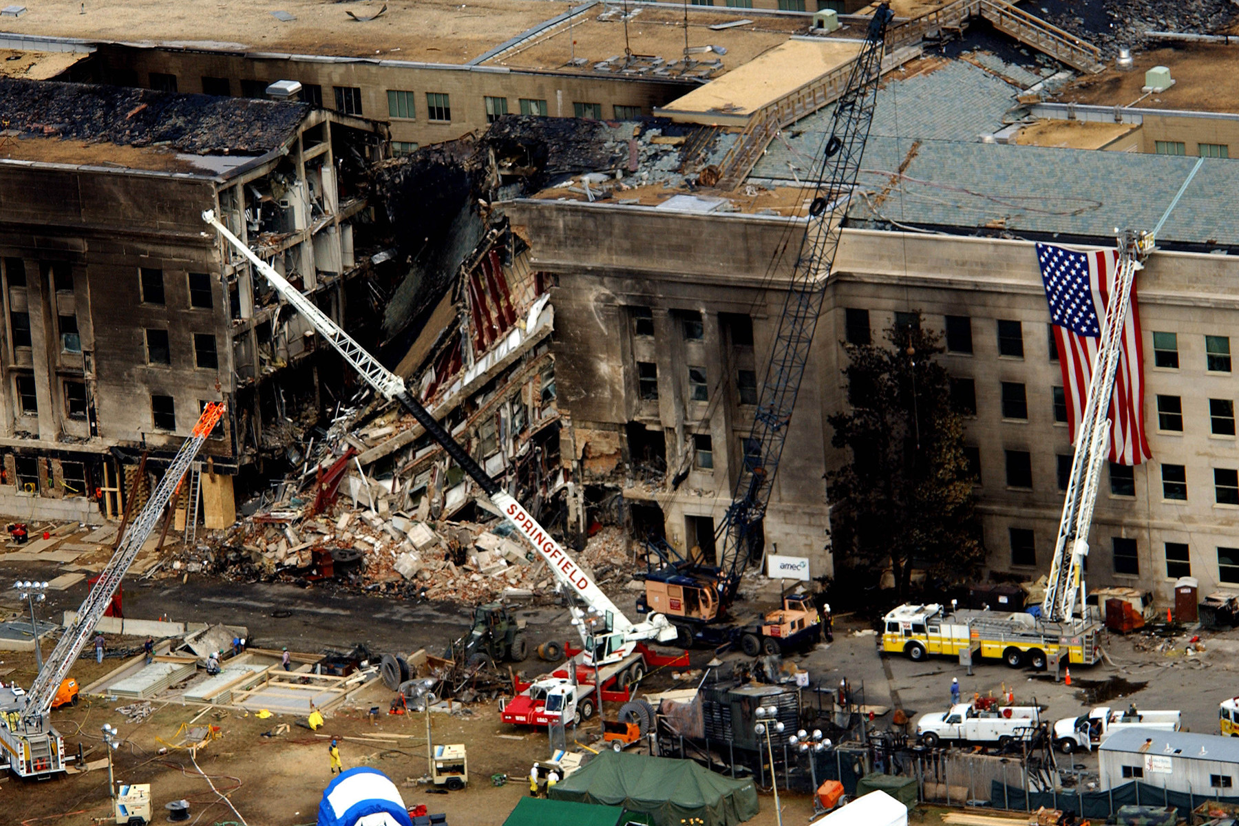 An aerial view of the west side of the Pentagon two days after the attack. A section of the outer rings has collapsed into a pile of rubble, the limestone facade above it is scorched black, and crane booms and fire ladders reach in from the lawn. A large American flag hangs from one of the ladders beside the damaged wall.