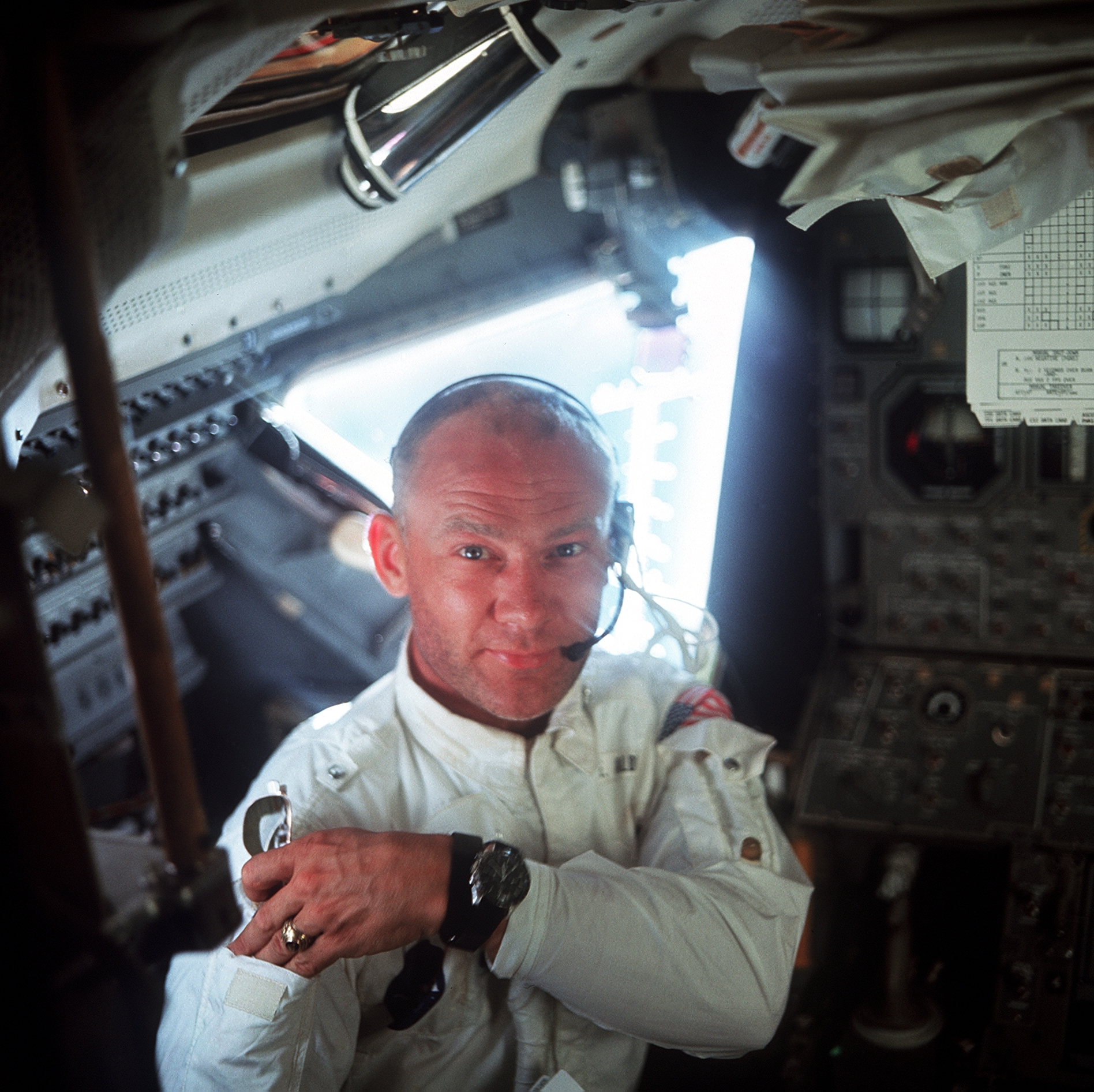 Buzz Aldrin inside the lunar module Eagle, wearing a headset, with the cabin's instrument panels and window behind him.